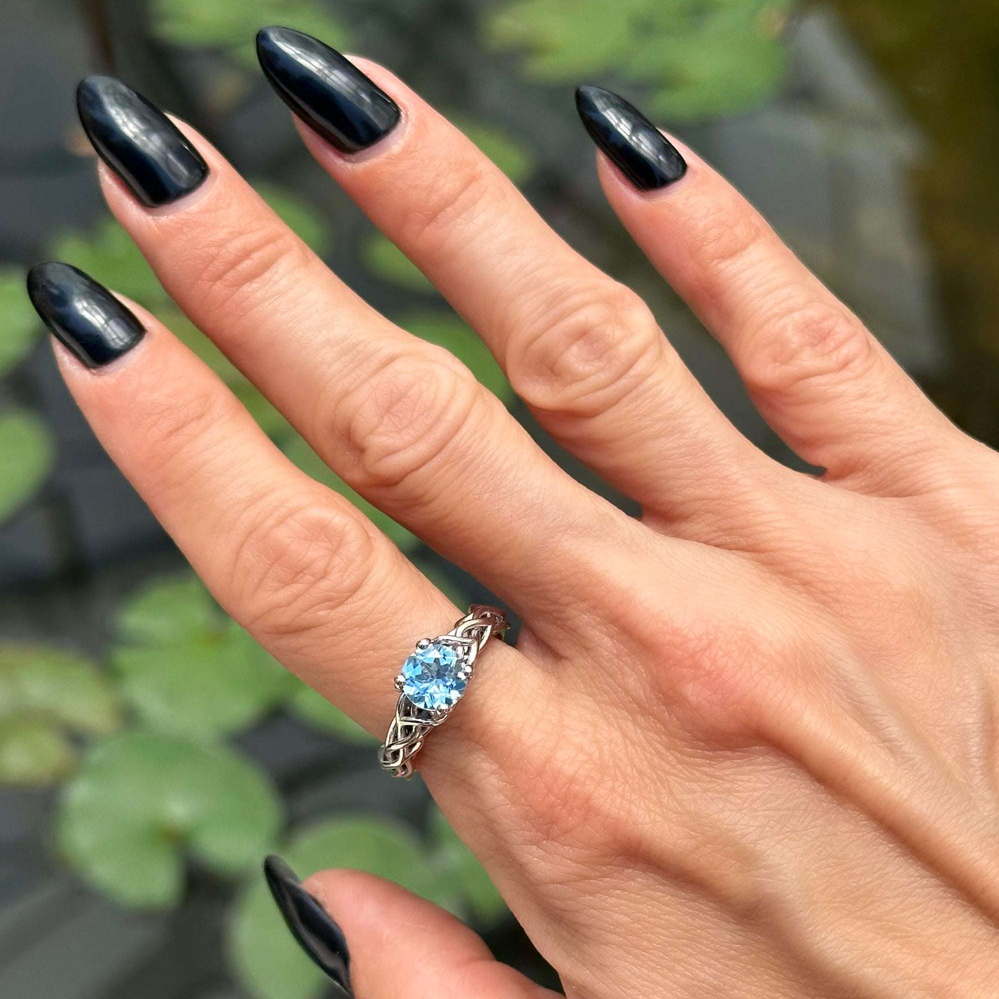 יד של אישה עם לק שחור שעונדת אבן חודש מרץ כחולה עם רקע ירוק