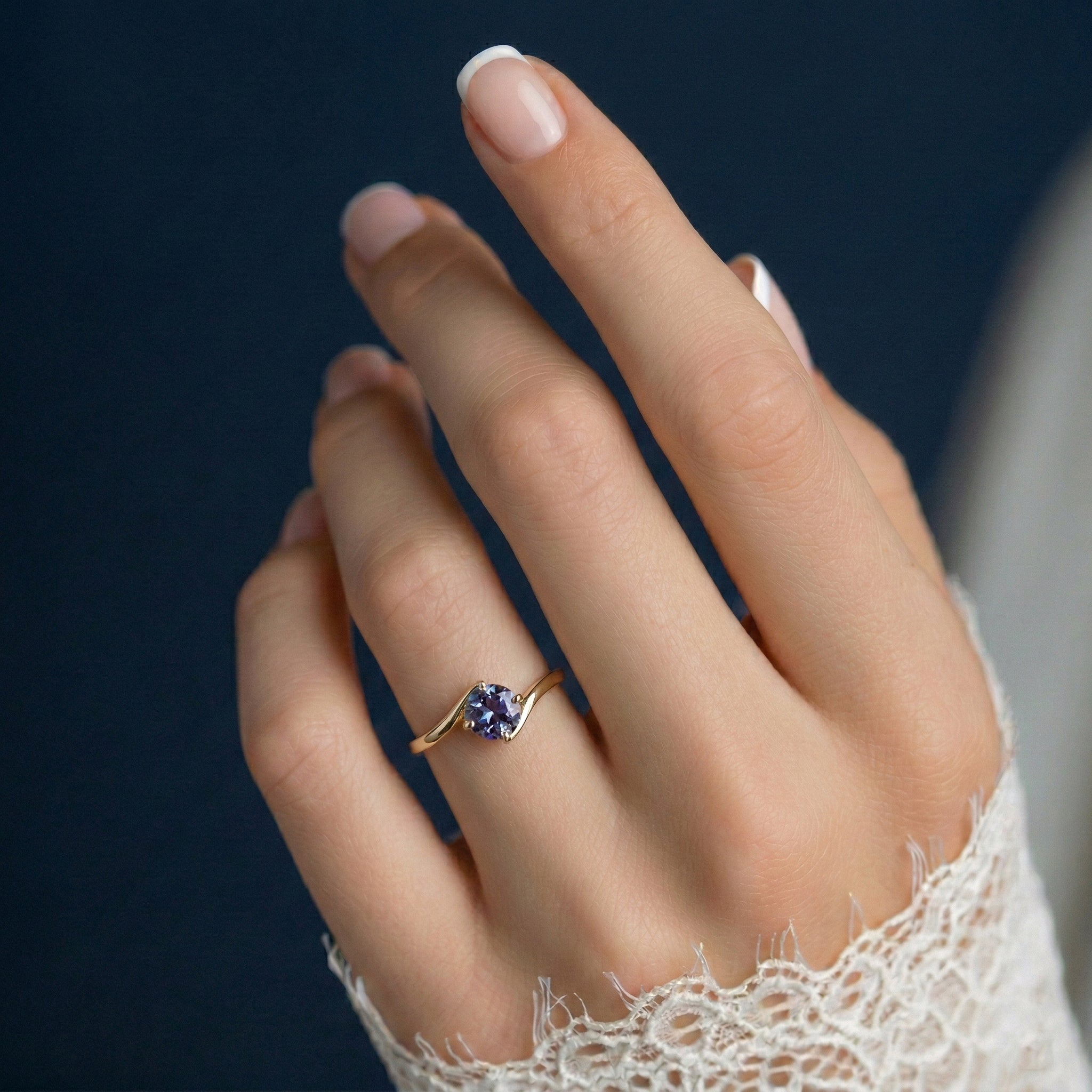 תקריב של יד של אישה שעונדת טבעת זהב צהוב משובצת אבן חודש יוני- אלכסנדריט על רקע כחול