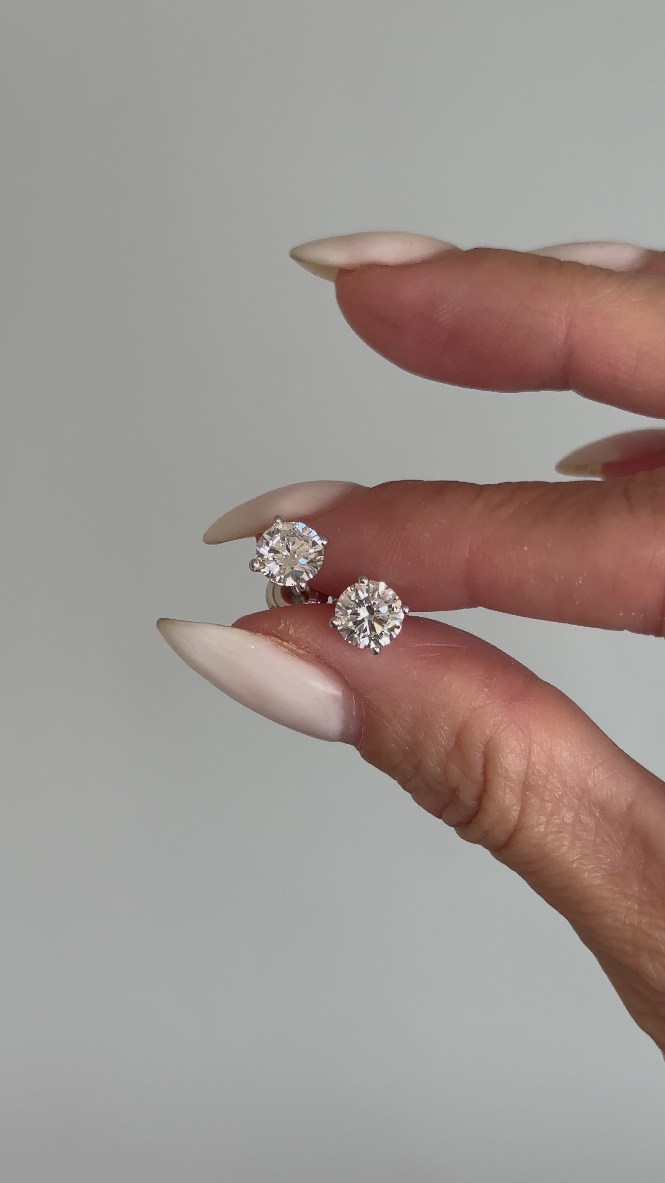 אצבעות של אישה שמחזיקות עגילי יהלומים צמודים בזהב לבן על רקע אפור